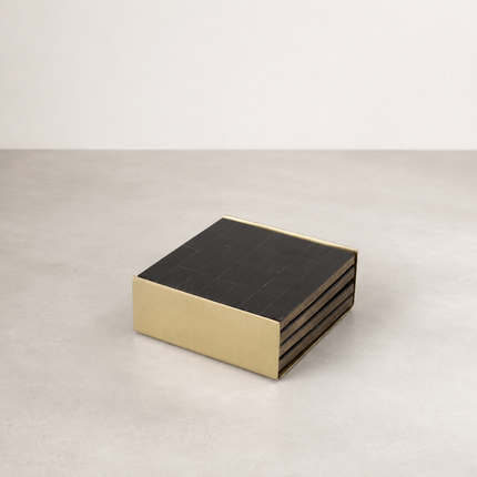 Black bone coasters stacked in a solid brass holder on a pale concrete surface