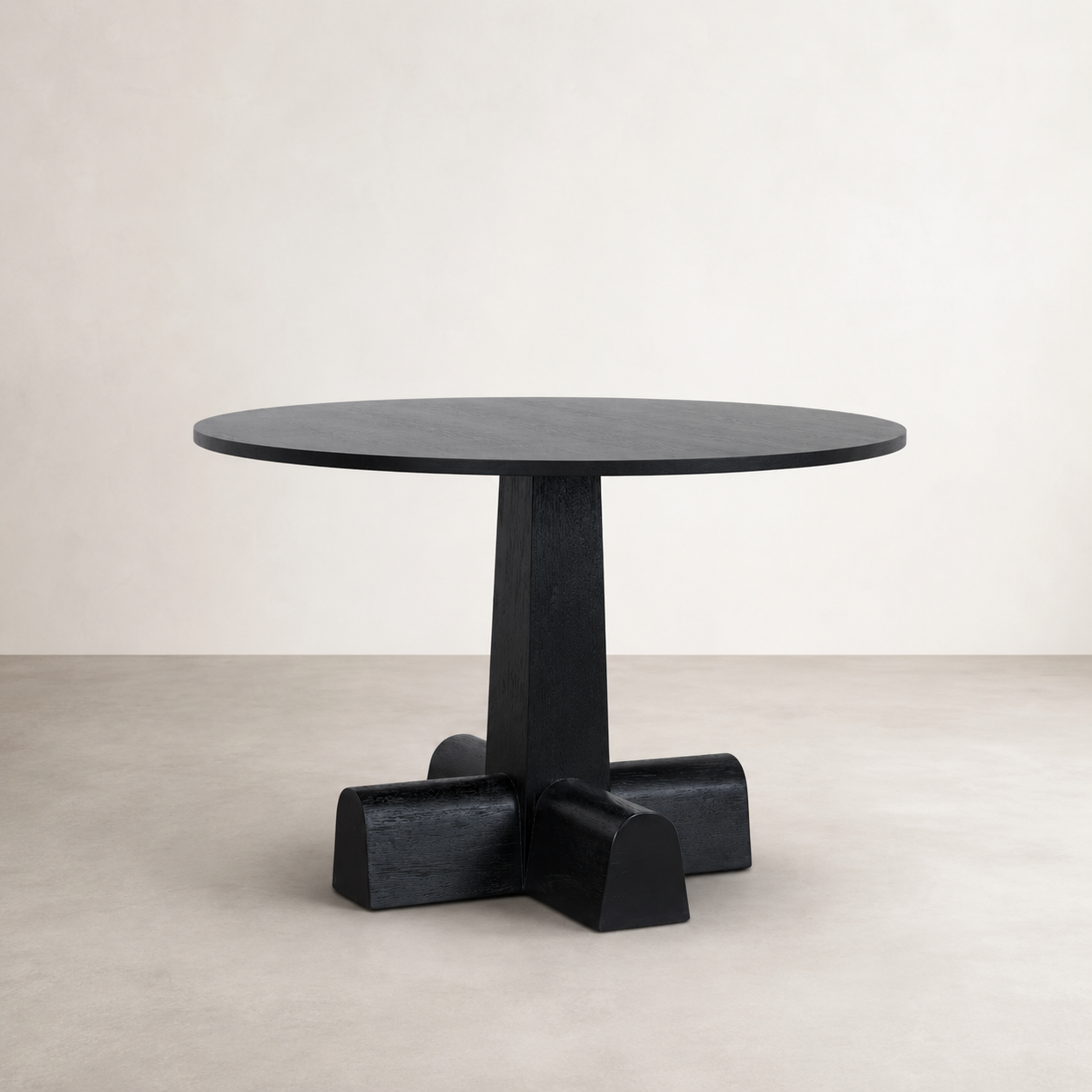 A round black dining table with a thick pedestal base and four block-like feet, photographed in a minimalist studio setting with a light beige wall and floor.