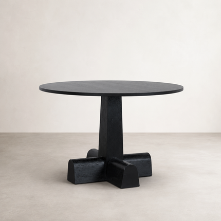 A round black dining table with a thick pedestal base and four block-like feet, photographed in a minimalist studio setting with a light beige wall and floor.