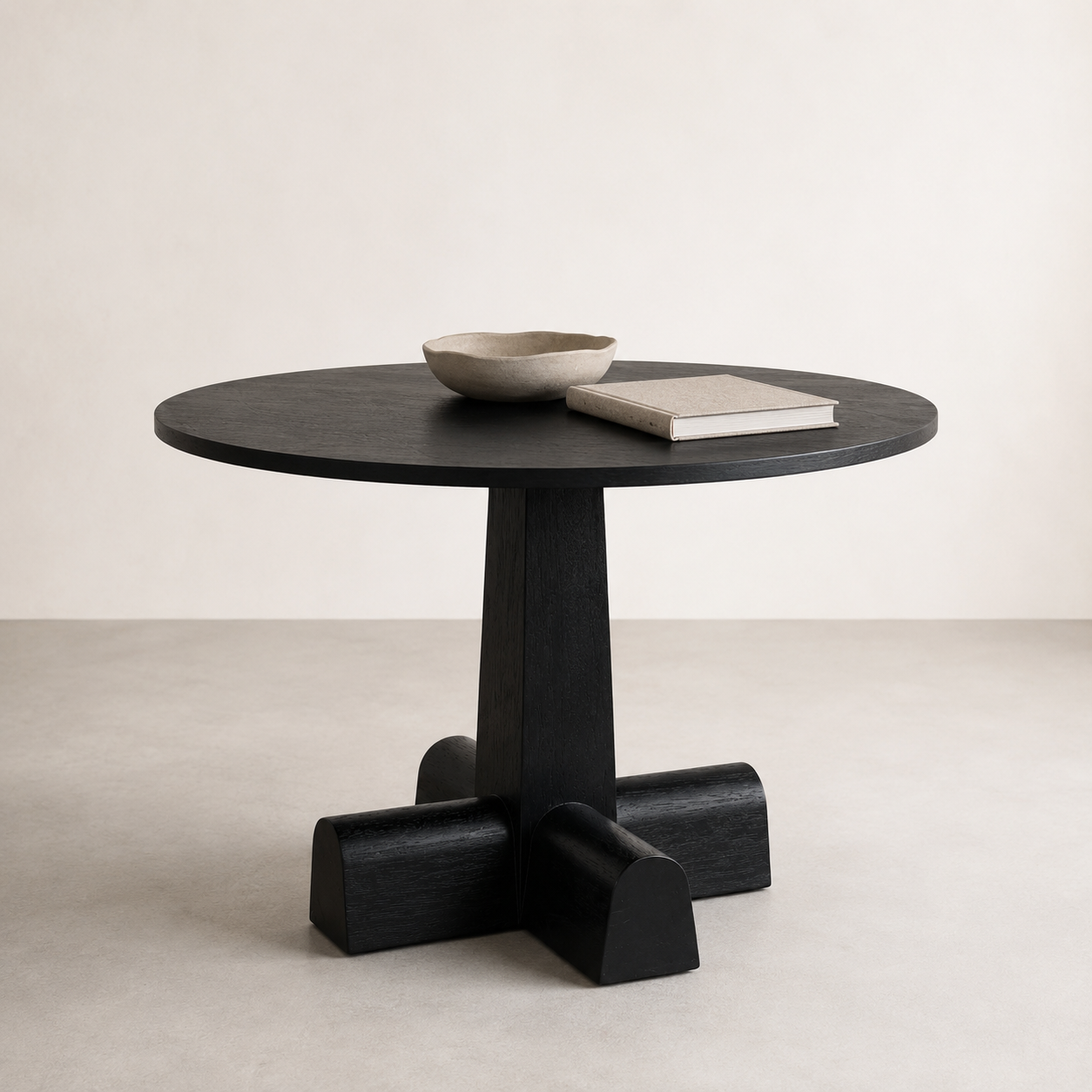 A round black wooden table with a thick pedestal base and four blocky legs, styled with a small bowl and a book on top, set against a minimalist beige background.