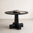 A round black wooden table with a thick pedestal base and four blocky legs, styled with a small bowl and a book on top, set against a minimalist beige background.