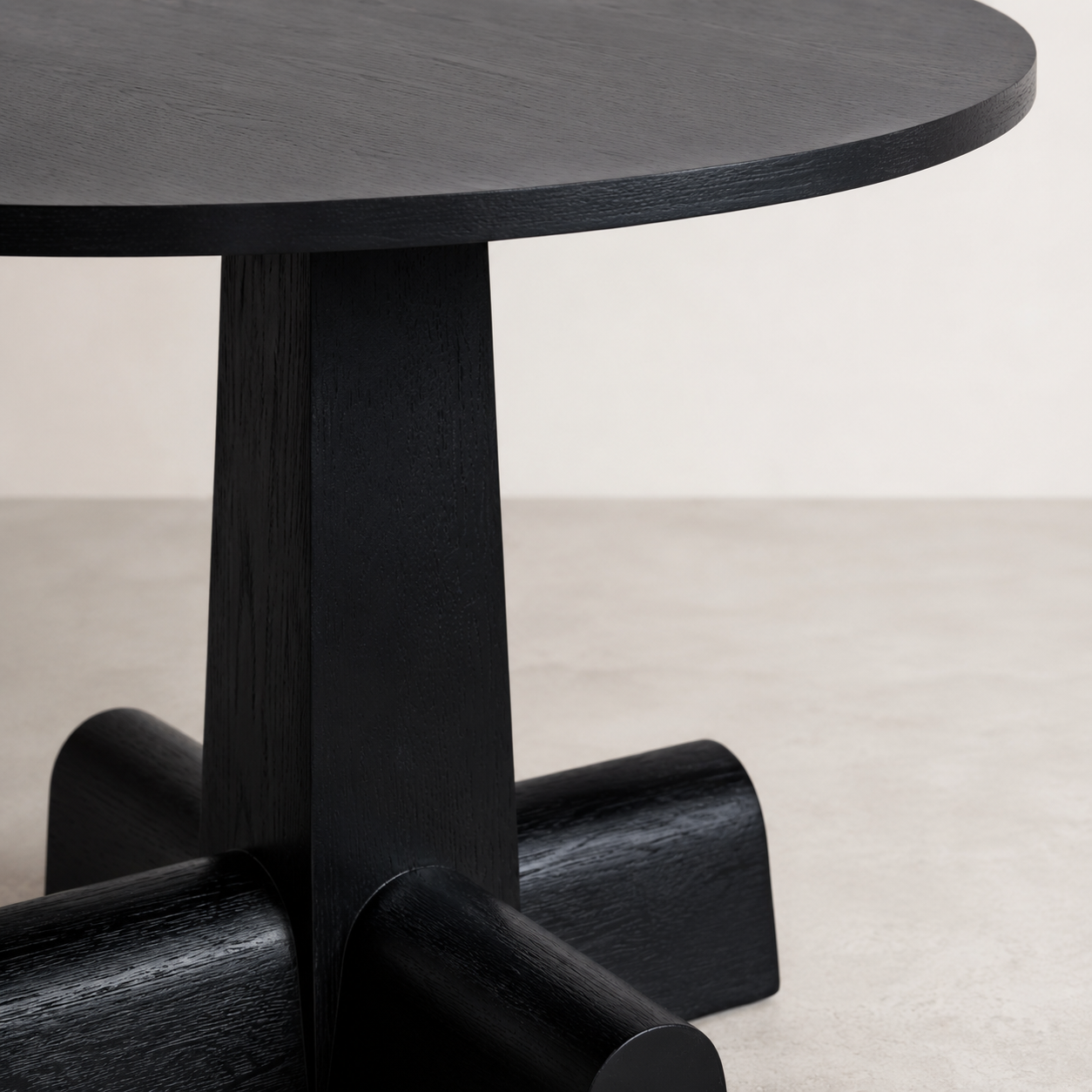Close-up of a black wooden table base and round tabletop against a light neutral wall and floor.