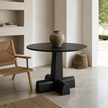 A round black dining table with a sculptural cross-shaped pedestal base, styled in a minimalist modern room with a woven chair and a ceramic vase on top.