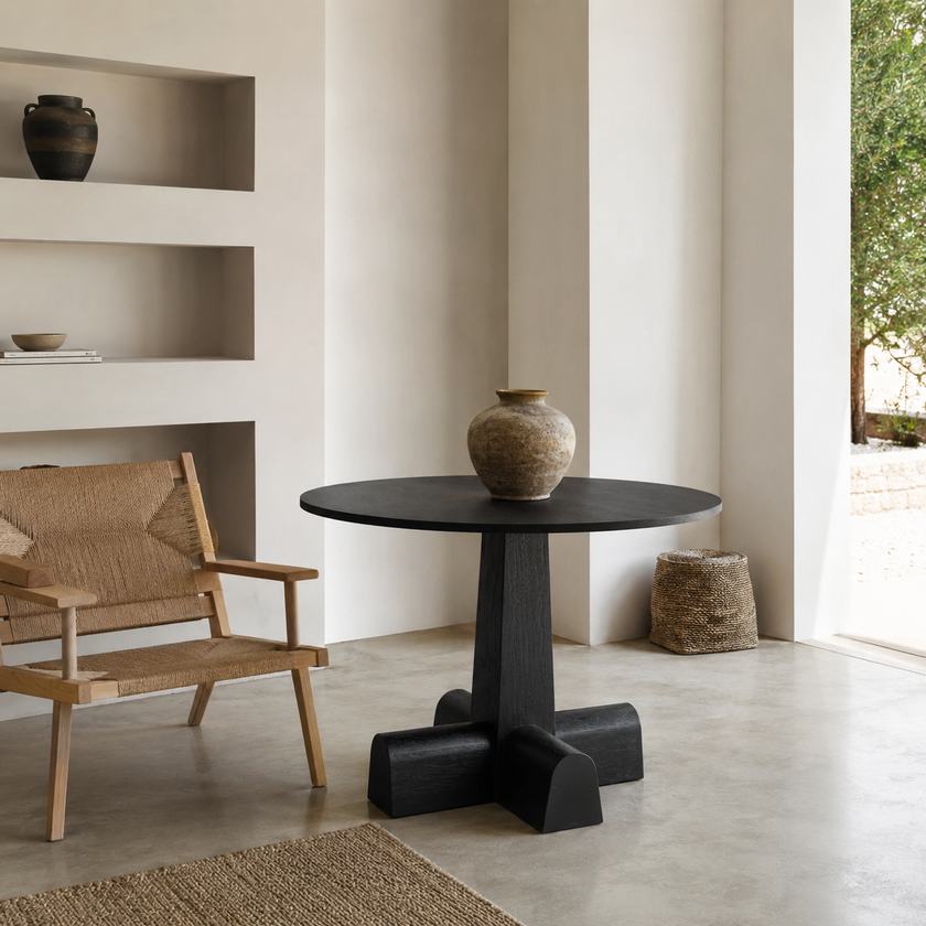 A round black pedestal table with a thick sculptural base stands in a minimalist interior room. A woven chair is to the left, and a textured vase sits on top of the table.