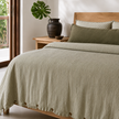 Olive striped French linen quilt cover over a timber bed beside a dark ceramic vase
